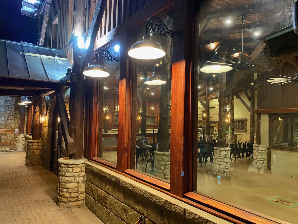 Covered porch of the timber-frame hall at night: pendant lights, timber posts on stone bases and tall wood-framed windows